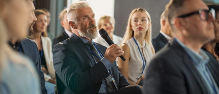 Senior Male Investor Sitting In Crowd And Using Microphone To Ask A Question At International Technology Conference. Caucasian Man Listening To Keynote Presentation About New Startup Company Service.